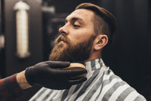Barber applying beard oil to a customer's beard with a striped apron and black gloves.