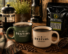 Two mugs with DESTIHL Brewery branding on a wooden table with coffee beans and a coffee bag.