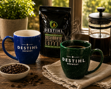 Two mugs with DESTIHL Brewery branding, a bag of coffee, and a French press on a wooden table.