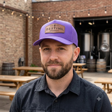 Man wearing a purple DESTIHL Brewery cap in front of a brick building with outdoor seating.