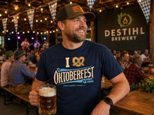 A man in a bar wearing a navy blue DESTIHL Brewery t-shirt with an Oktoberfest graphic, holding a glass of beer.