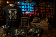 A dark-themed retail display featuring a black DESTIHL Brewery graphic T-shirt with a skull-and-beer-mug design, along with matching folded T-shirts, a branded wooden crate, a cap, and glasses, set in a dimly lit environment.