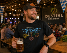 A man smiling in a brewery holding a beer mug, wearing a black DESTIHL Brewery t-shirt with a colorful Oktoberfest graphic and a dark baseball cap.